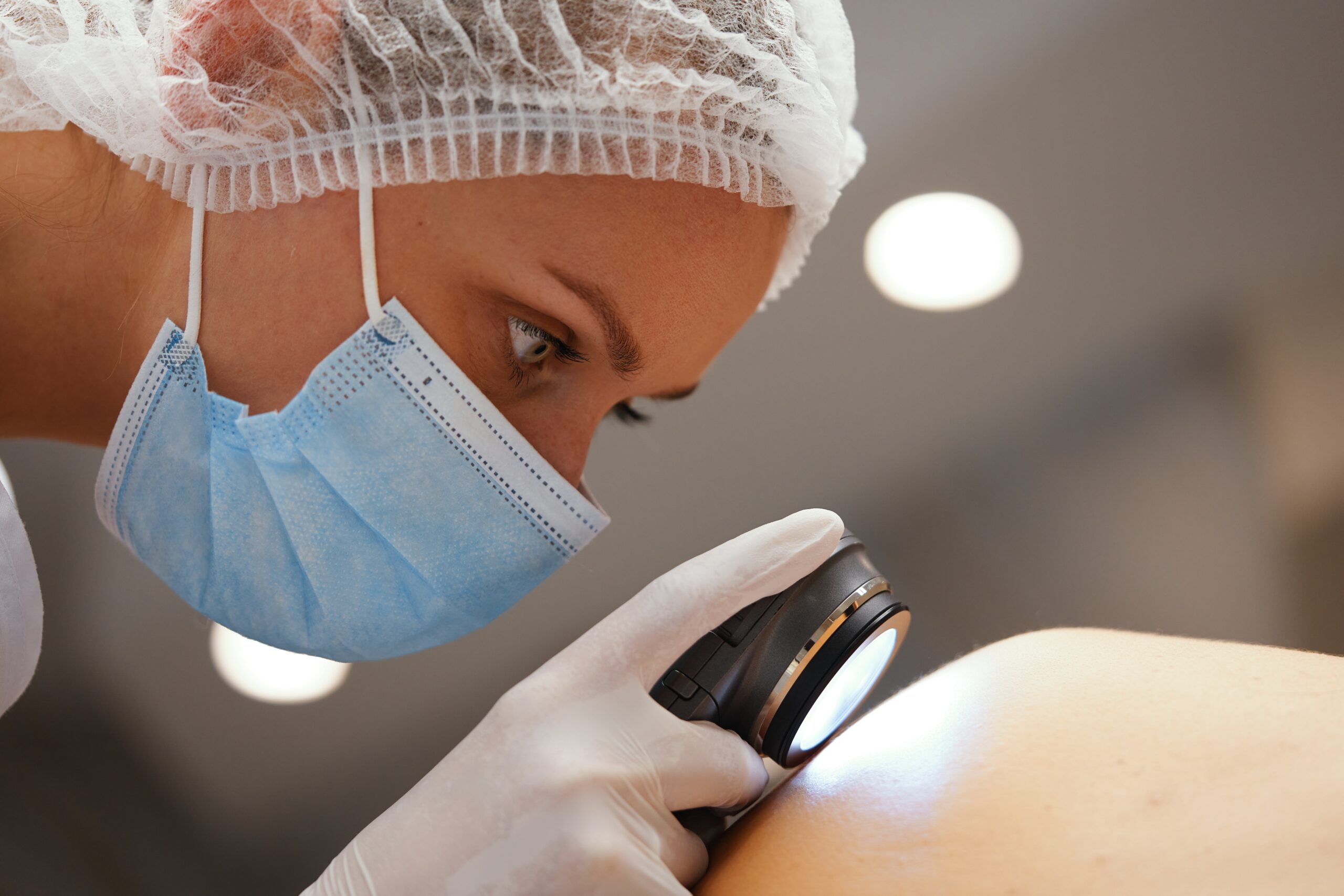 Close-up of a clinician wearing a disposable cap and surgical mask, looking into a surgical microscope during a procedure with gloved hands visible