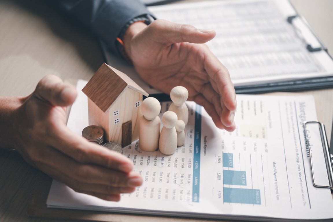Hands cradle a wooden house model with small figurines and coins on a mortgage document.
