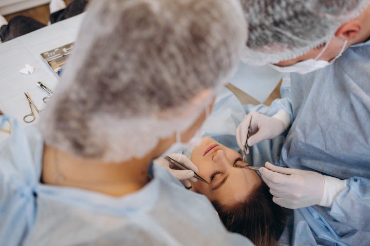 Two dental professionals in scrubs and masks treat a patient lying in a chair, using dental instruments near the mouthבס.