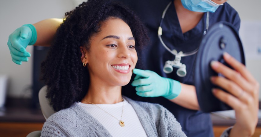 Smiling woman in a dental chair as a clinician in gloves and a mask checks her teeth with a mirror tool?
