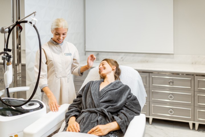 Clinical aesthetic treatment: clinician in a beige uniform stands beside a smiling patient in a gray robe on a treatment chair with nearby equipment.