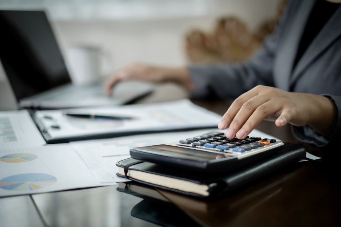 Hands typing on a calculator atop a notebook with financial charts and a laptop in the background.