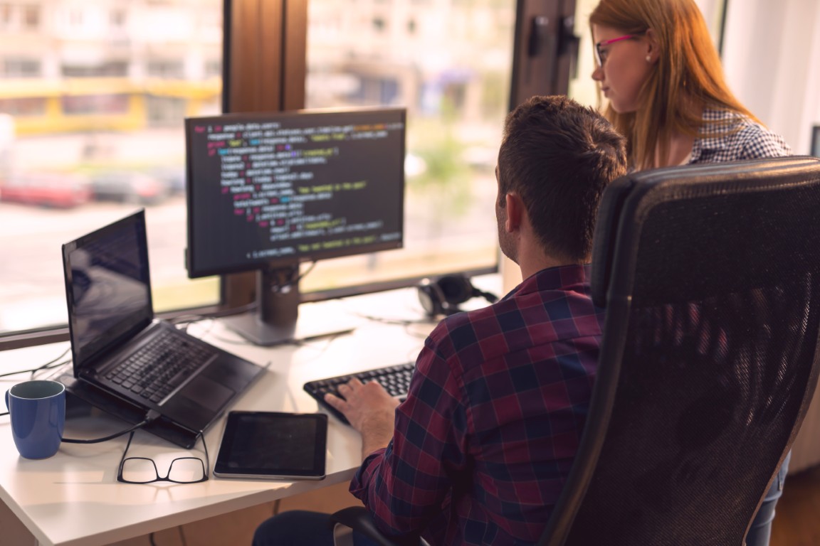 Two programmers work at a desk: man typing at a keyboard while a woman looks on, with code on a large monitor and laptops nearby.