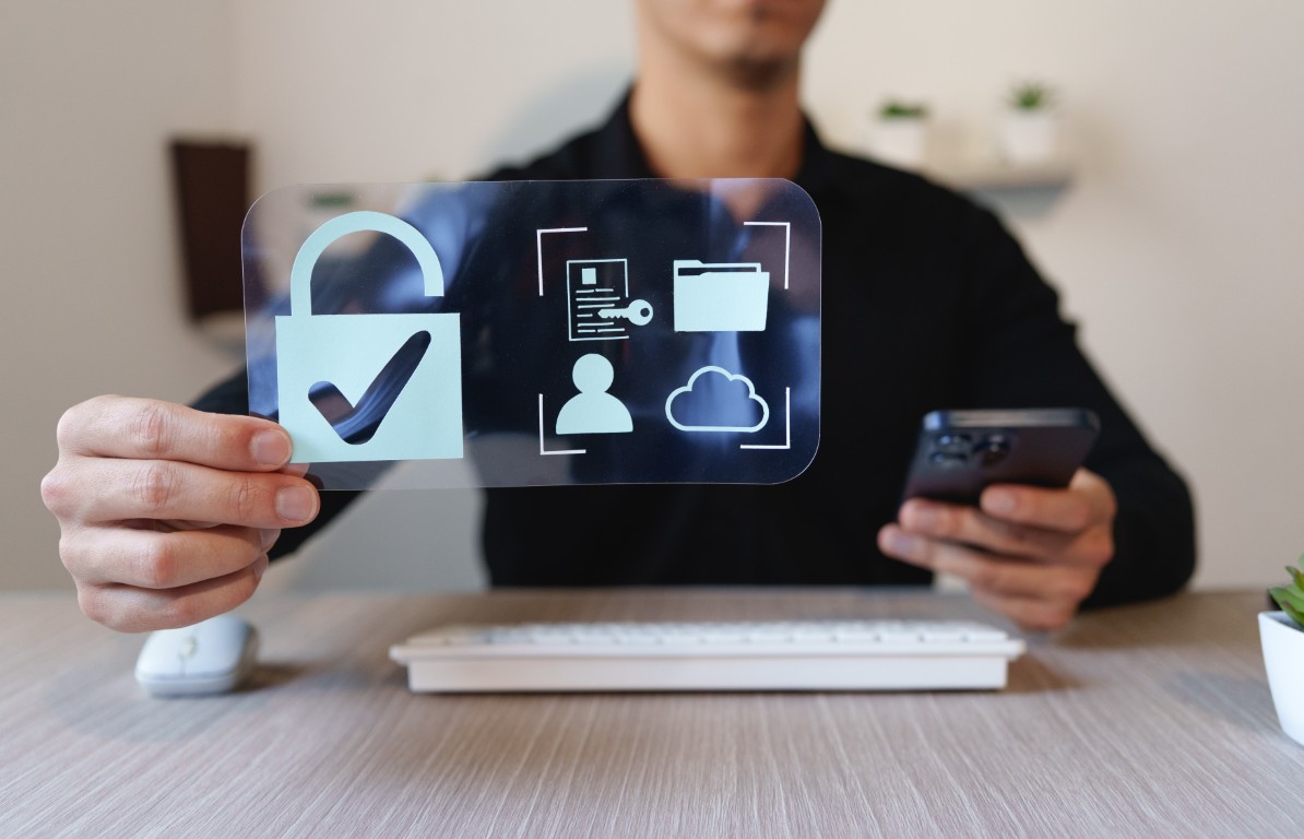 Person at a desk holds a translucent card showing a large padlock and data-security icons (user, cloud, folder) while looking at a smartphone.