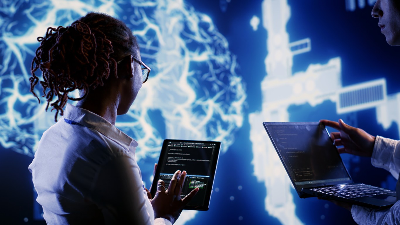 Two professionals collaborate, one on a tablet with code and one on a laptop, in a high-tech blue-lit setting.