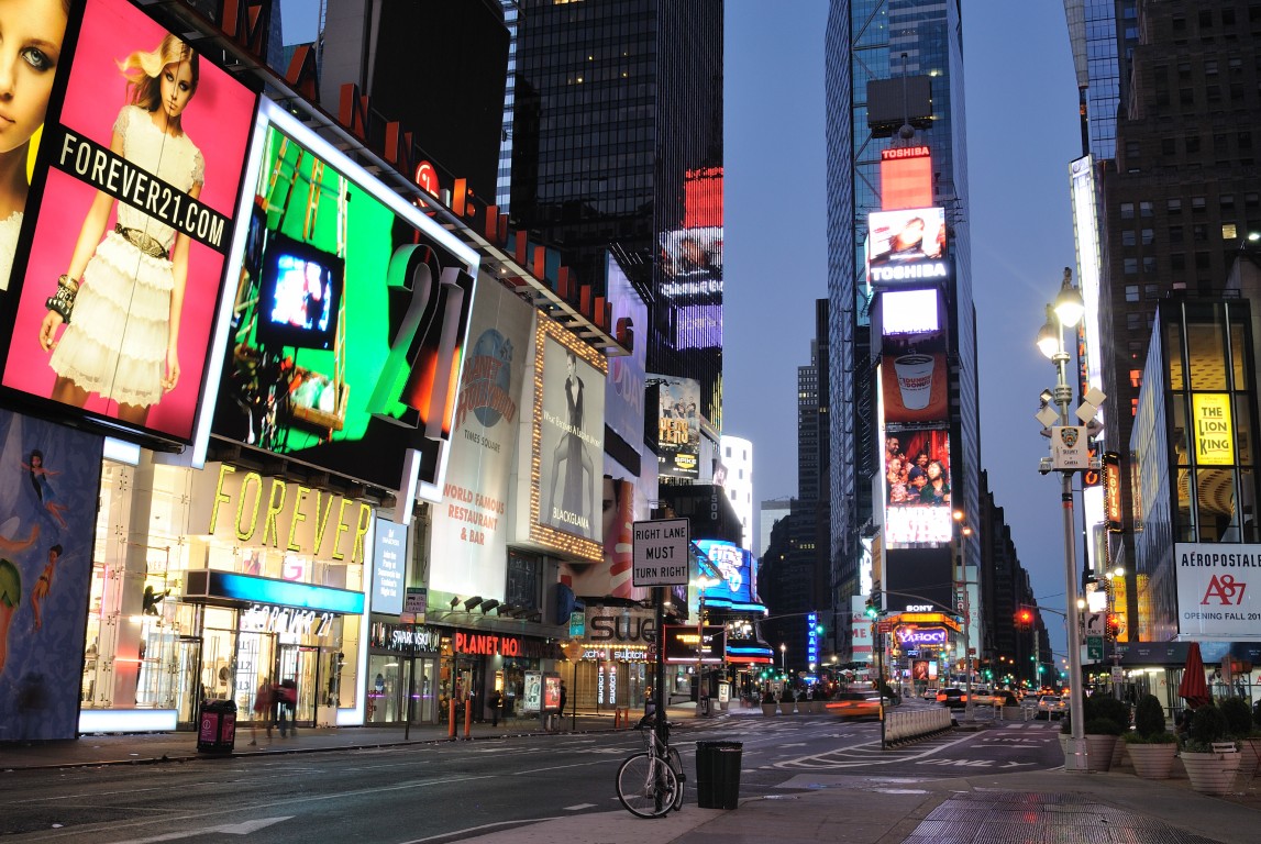 Bright Times Square with large colorful billboards and neon signs at dusk, tall buildings framing the street.